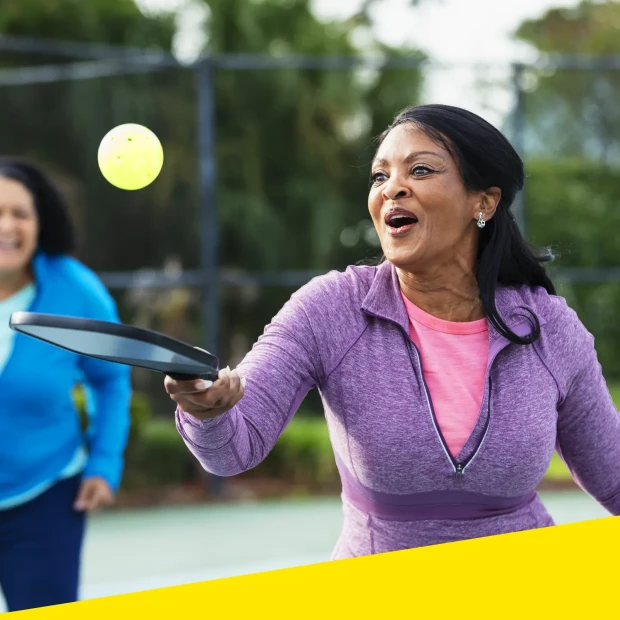 A woman smiles while bouncing a pickleball on a paddle with friends.